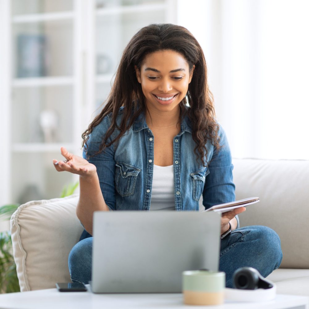 A woman sits comfortably on a sofa, interacting with her laptop while smiling. She is actively participating in a virtual meeting with her tablet close by, showcasing a cozy home environment.